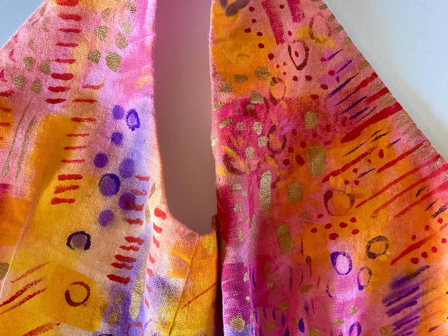 A piece of handpainted clothing. It is in yellow/orange/red/purple/pink washes with purple/red/gold abstract detailing in lines, circles and dots. 