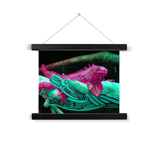 A black wooden poster hanger. A digital collage: a photo of an Iguana laying on a branch, doodles are rendered on the Iguana and the branch. The iguana is pink and the branch is seafoam green. 