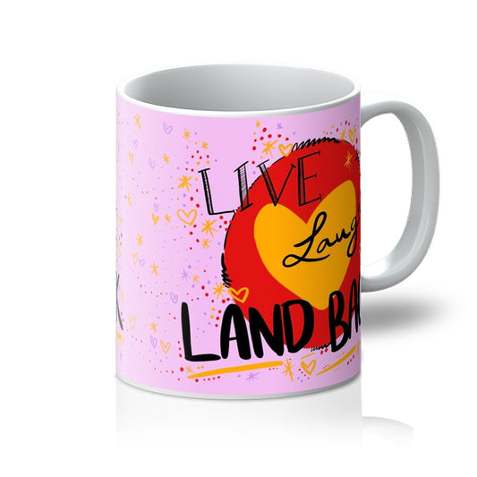White mug printed with art. ‘live laugh land back’ written in black with a yellow heart and red circle behind. The background is pink with small dots, squiggles, stars and hearts dotted around the image in red, yellow and purple. Handle is on the right. 