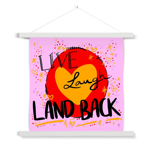 White wooden poster hanger. The art print is square. The design: ‘live laugh land back’ written in black with a yellow heart and red circle behind. The background is pink with small dots, squiggles, stars and hearts dotted around the image in red, yellow and purple.