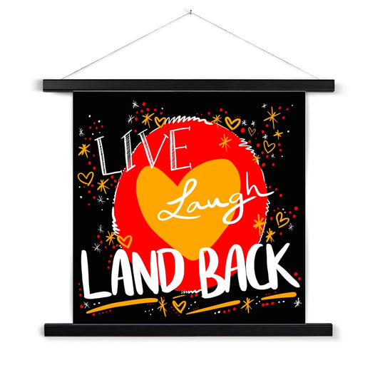 A black wooden poster hanger. The print is square. The design: ‘live laugh land back’ written in white with a yellow heart and red circle behind. The background is black with small dots, squiggles, stars and hearts dotted around the image in red, yellow and white.
