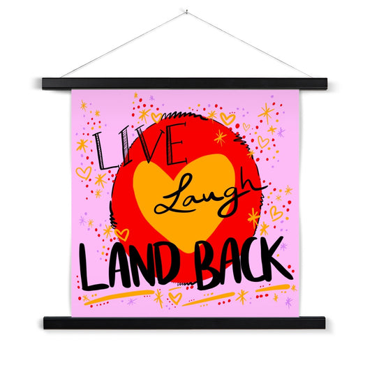Black wooden poster hanger. The art print is square. The design: ‘live laugh land back’ written in black with a yellow heart and red circle behind. The background is pink with small dots, squiggles, stars and hearts dotted around the image in red, yellow and purple.